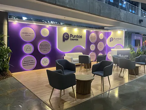 Stand ferial de Puntos Colombia en el Inbound Summit 2026, con paneles luminosos morados, mesas de madera y piso de madera clara