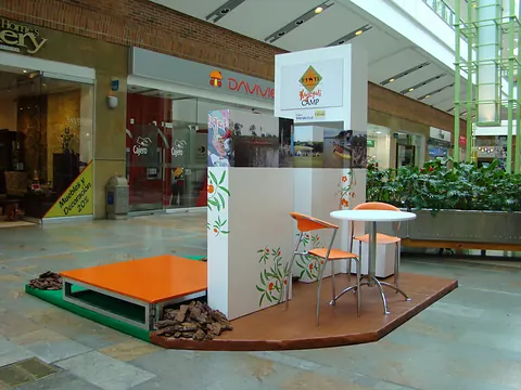 Stand comercial en un centro comercial, con plataforma naranja, mesa alta y panel de marca