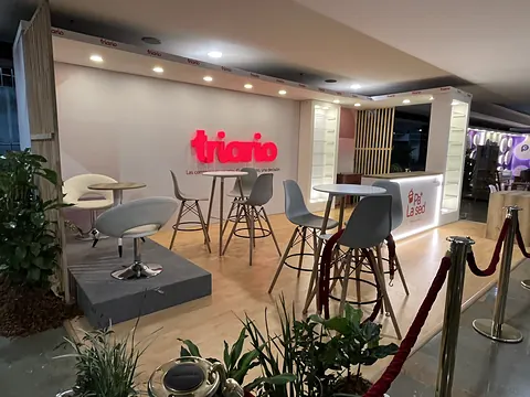 Stand de Triario en el Inbound Summit 2026 con mostrador iluminado, mesas altas, banquetas, cordones de protocolo y plantas